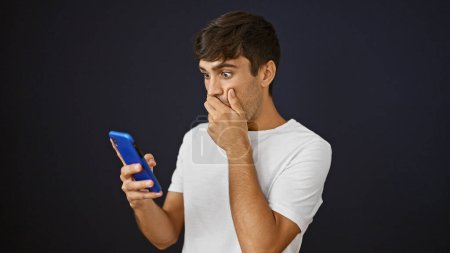 Foto de Hombre hispano joven mirando el teléfono inteligente con expresión sorpresa sobre un fondo negro aislado - Imagen libre de derechos