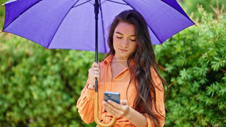 Téléchargez les photos : Jeune belle femme hispanique tenant parapluie en utilisant un smartphone au parc - en image libre de droit