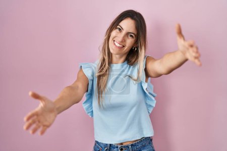 Téléchargez les photos : Jeune femme hispanique debout sur fond rose regardant la caméra souriant à bras ouverts pour un câlin. expression joyeuse embrassant le bonheur. - en image libre de droit