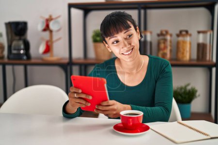 Téléchargez les photos : Jeune belle femme hispanique utilisant touchpad boire du café assis sur la table à la maison - en image libre de droit