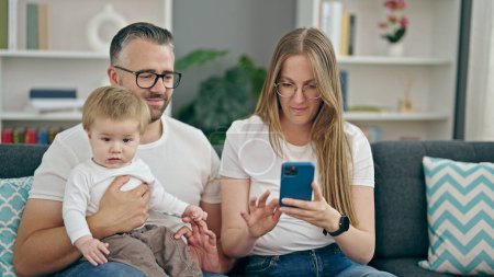 Photo for Family of mother, father and baby smiling using smartphone sitting on the sofa at home - Royalty Free Image