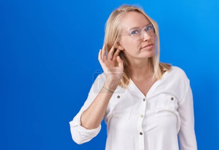 Téléchargez les photos : Young caucasian woman standing over blue background smiling with hand over ear listening an hearing to rumor or gossip. deafness concept. - en image libre de droit