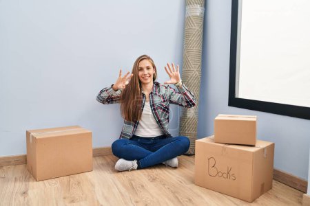 Photo for Young blonde woman sitting on the floor moving to a new home showing and pointing up with fingers number eight while smiling confident and happy. - Royalty Free Image