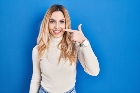 Photo for Young caucasian woman standing over blue background pointing with hand finger to face and nose, smiling cheerful. beauty concept - Royalty Free Image