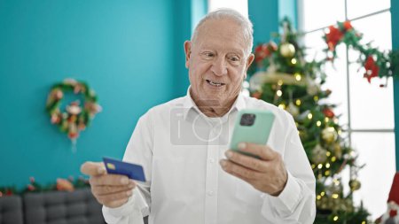 Foto de Hombre mayor de pelo gris de compras con teléfono inteligente y tarjeta de crédito celebrando la Navidad en casa - Imagen libre de derechos