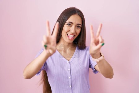 Foto de Mujer hispana joven con el pelo largo de pie sobre fondo rosa sonriendo con la lengua hacia fuera mostrando los dedos de ambas manos haciendo signo de victoria. número dos. - Imagen libre de derechos