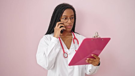 Foto de Mujer afroamericana doctora leyendo documento en portapapeles hablando en smartphone sobre fondo rosa aislado - Imagen libre de derechos