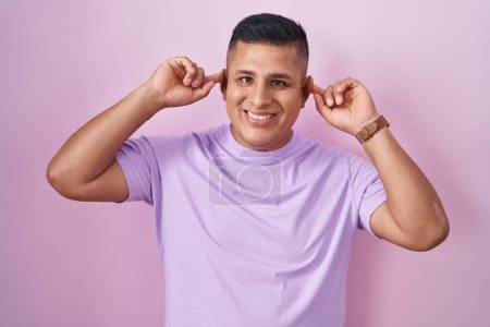 Foto de Joven hombre hispano de pie sobre fondo rosa sonriendo tirando de las orejas con los dedos, gesto divertido. problema de audición - Imagen libre de derechos