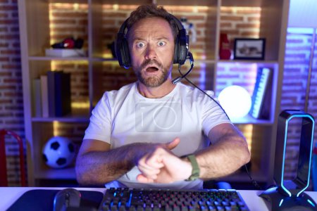 Téléchargez les photos : Homme du Moyen Âge avec barbe jouer à des jeux vidéo portant des écouteurs à la hâte pointant vers regarder le temps, l'impatience, bouleversé et en colère pour retard de délai - en image libre de droit