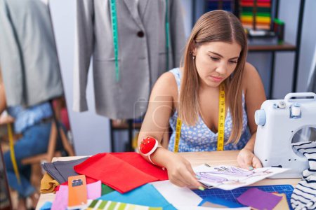 Téléchargez les photos : Jeune belle femme hispanique tailleur regardant la conception de vêtements au magasin de tailleur - en image libre de droit