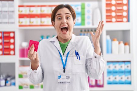 Téléchargez les photos : Femme brune travaillant à la pharmacie pharmacie tenant préservatif célébrant la victoire avec sourire heureux et expression gagnante avec les mains levées - en image libre de droit