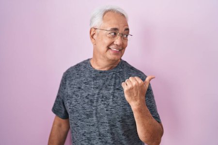 Photo for Middle age man with grey hair standing over pink background smiling with happy face looking and pointing to the side with thumb up. - Royalty Free Image