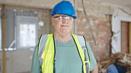 Foto de Constructor de hombre de pelo gris de mediana edad sonriendo confiado de pie en el sitio de construcción - Imagen libre de derechos