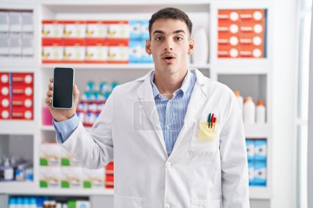 Foto de Hombre hispano guapo trabajando en farmacia mostrando pantalla de teléfono inteligente asustado y sorprendido con la boca abierta para sorpresa, cara de incredulidad - Imagen libre de derechos
