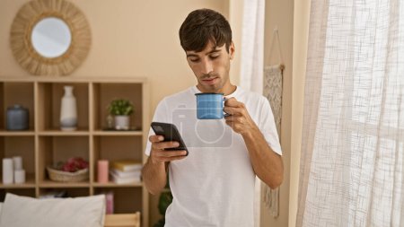 Photo for Handsome young hispanic man quietly enjoying his morning coffee at home, fully immersed in texting on his smartphone, comfortably seated in his living room, basking under the warm indoor lighting - Royalty Free Image