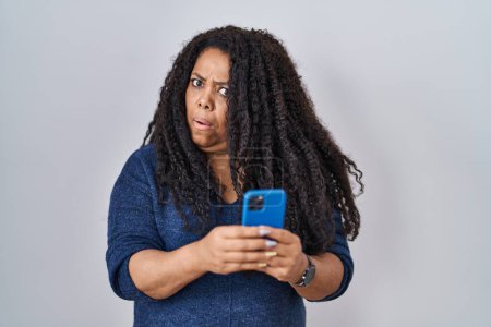 Téléchargez les photos : Plus taille femme hispanique en utilisant un message de dactylographie smartphone dans le visage de choc, l'air sceptique et sarcastique, surpris avec la bouche ouverte - en image libre de droit