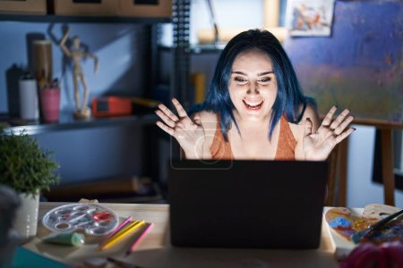 Téléchargez les photos : Jeune fille moderne avec les cheveux bleus assis au studio d'art avec ordinateur portable la nuit célébrant fou et étonné pour le succès avec les bras levés et les yeux ouverts hurlant excités. concept gagnant - en image libre de droit