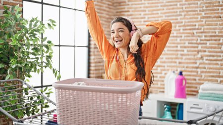 Foto de Joven mujer hispana hermosa escuchando música colgando ropa en el tendedero bailando en la lavandería - Imagen libre de derechos