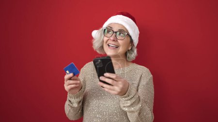 Téléchargez les photos : Femme du Moyen Age avec les cheveux gris faire des achats en ligne de Noël avec smartphone sur fond rouge isolé - en image libre de droit