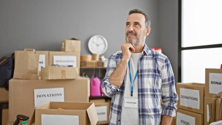 Photo for Serious middle age man, with grey hair and beard, a thoughtful volunteer at charity center, doubting over donations in a cardboard box - Royalty Free Image