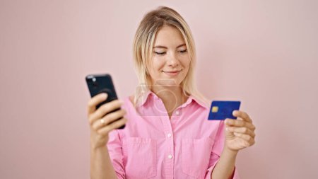 Téléchargez les photos : Jeune femme blonde faisant du shopping en ligne avec smartphone et carte de crédit sur fond rose isolé - en image libre de droit