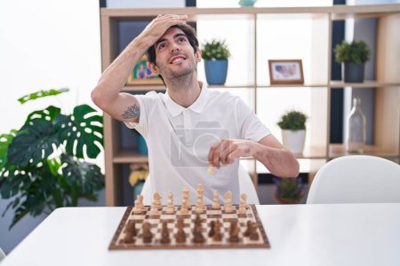 Téléchargez les photos : Jeune homme hispanique jouant aux échecs assis sur la table stressé et frustré avec la main sur la tête, visage surpris et en colère - en image libre de droit