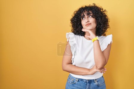 Téléchargez les photos : Jeune femme du Moyen-Orient debout sur fond jaune avec la main sur le menton penser à la question, expression coûteuse. souriant avec un visage réfléchi. concept de doute. - en image libre de droit