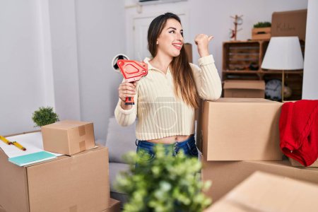 Téléchargez les photos : Jeune femme hispanique se déplaçant vers une nouvelle maison boîtes d'emballage pointant pouce vers le côté souriant heureux avec bouche ouverte - en image libre de droit