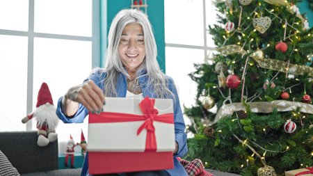 Foto de Mujer de pelo gris de mediana edad desempacando regalo de Navidad sonriendo confiado en casa - Imagen libre de derechos
