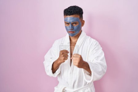 Foto de Joven hombre hispano usando mascarilla de belleza y bata de baño listo para luchar con el gesto de defensa del puño, cara enojada y molesta, miedo al problema - Imagen libre de derechos