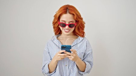 Téléchargez les photos : Jeune rousse femme portant des lunettes de soleil en utilisant smartphone sur fond blanc isolé - en image libre de droit