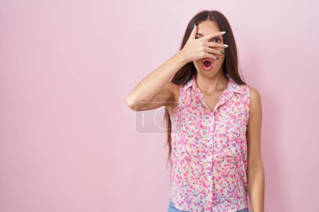 Foto de Mujer hispana joven con el pelo largo de pie sobre el fondo rosa asomándose en shock cubriendo la cara y los ojos con la mano, mirando a través de los dedos con expresión avergonzada. - Imagen libre de derechos