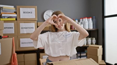 Téléchargez les photos : Jeune femme blonde bénévole souriant confiant faire geste de coeur au centre de charité - en image libre de droit