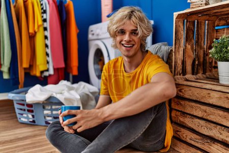 Photo for Young blond man drinking coffee waiting for washing machine at laundry room - Royalty Free Image
