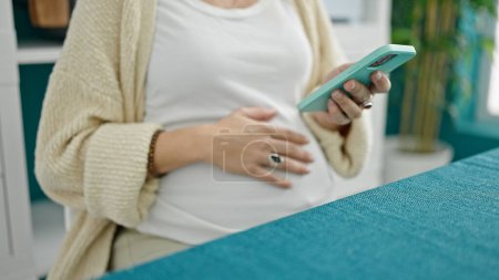 Photo for Young pregnant woman using smartphone touching belly at dinning room - Royalty Free Image