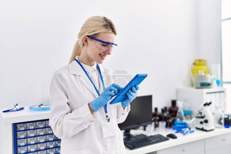 Téléchargez les photos : Jeune femme blonde scientifique utilisant pavé tactile souriant au laboratoire - en image libre de droit