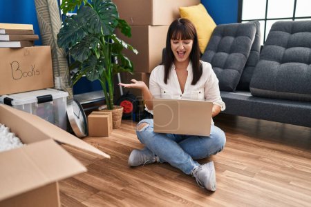 Téléchargez les photos : Jeune femme brune assise sur le sol avec un ordinateur portable à la nouvelle maison célébrant la réalisation avec un sourire heureux et l'expression gagnante avec la main levée - en image libre de droit