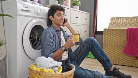 Téléchargez les photos : Jeune homme hispanique parlant sur smartphone boire du café en attendant la machine à laver à la buanderie - en image libre de droit
