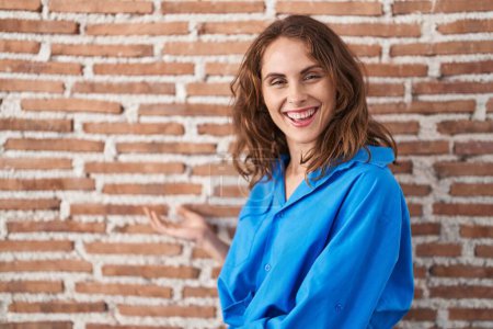 Foto de Hermosa mujer morena de pie sobre la pared de ladrillos invitando a entrar sonriendo natural con la mano abierta - Imagen libre de derechos