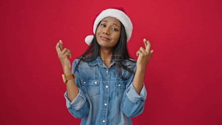 Foto de Mujer afroamericana con sombrero de navidad cruzando los dedos a la suerte sobre fondo rojo aislado - Imagen libre de derechos
