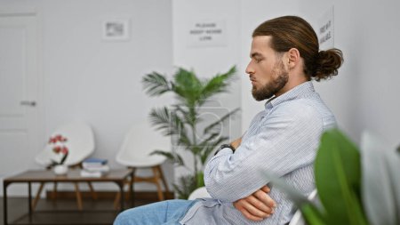 Téléchargez les photos : Jeune homme hispanique assis sur une chaise avec des bras croisés geste et visage sérieux dans la salle d'attente - en image libre de droit