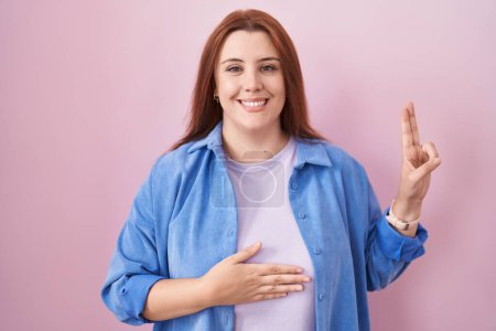 Téléchargez les photos : Jeune femme hispanique avec les cheveux roux debout sur fond rose en jurant souriant avec la main sur la poitrine et les doigts vers le haut, faisant un serment de fidélité - en image libre de droit