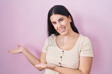 Photo for Young hispanic woman standing over pink background inviting to enter smiling natural with open hand - Royalty Free Image