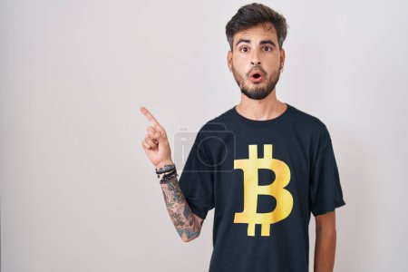 Photo for Young hispanic man with tattoos wearing bitcoin t shirt surprised pointing with finger to the side, open mouth amazed expression. - Royalty Free Image