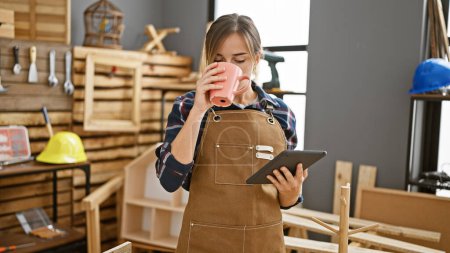 Foto de Atractiva joven carpintera rubia, café en la mano, navegando por el touchpad en medio del ajetreo de la carpintería en el animado taller de carpintería - Imagen libre de derechos