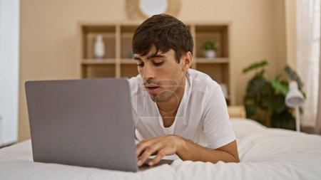 Foto de Guapo joven hispano descansando cómodamente en una cama acogedora, absorto en su computadora portátil, disfrutando de una mañana relajante en su dormitorio en casa. - Imagen libre de derechos