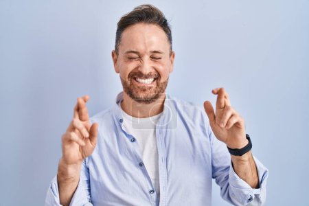Téléchargez les photos : Homme caucasien du Moyen Age debout sur fond bleu geste doigt croisé souriant avec espoir et les yeux fermés. chance et concept superstitieux. - en image libre de droit