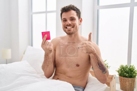 Téléchargez les photos : Jeune homme hispanique tenant préservatif dans le lit souriant heureux et positif, pouce levé faire excellent et signe d'approbation - en image libre de droit