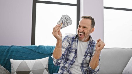 Foto de Hombre de mediana edad con pelo gris contando dólares sentado en el sofá celebrando en casa - Imagen libre de derechos