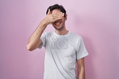 Foto de Joven hombre hispano de pie sobre fondo rosa sonriendo y riendo con la mano en la cara cubriendo los ojos para sorpresa. concepto ciego. - Imagen libre de derechos